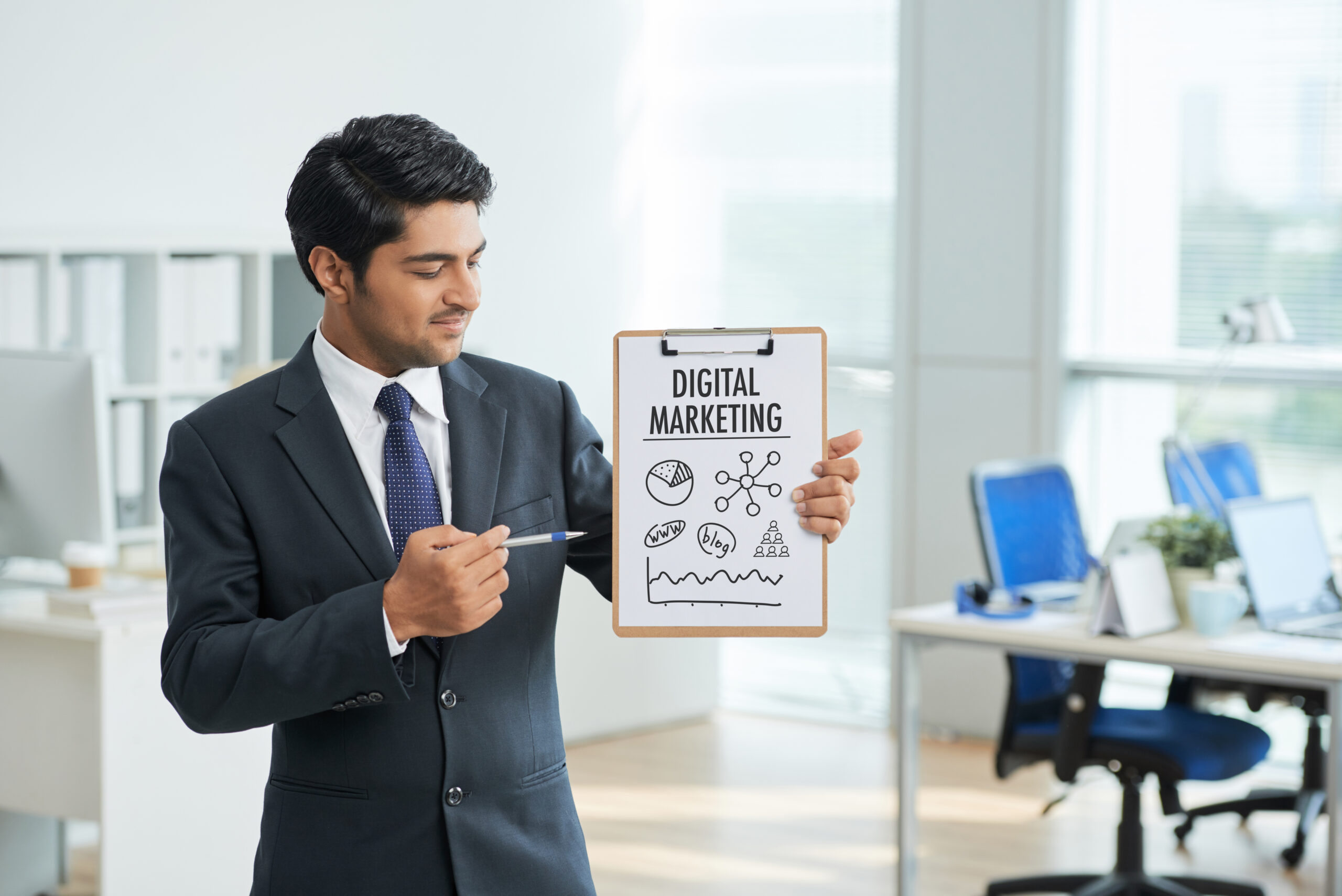A businessman in a suit stands in an office, holding a clipboard with a hand-drawn digital marketing concept, including icons for websites, blogs, and graphs.